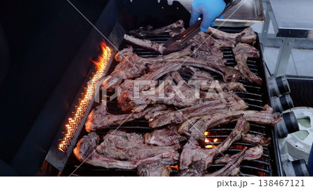 Chef wearing blue gloves uses tongs to grill lamb chops on a barbecue grill during an outdoor barbecue party, flames licking the meat as it cooks Chef wearing blue gloves uses tongs to grill lamb chops on a barbecue grill during an outdoor barbecue party, flames licking the meat as it cooks 138467121