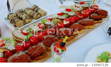 Rustic wooden tray displaying gourmet appetizers, including mini sandwiches, grilled eggplant rolls, cherry tomatoes, and charcuterie spread on crisp white linen at catered gathering 138467130