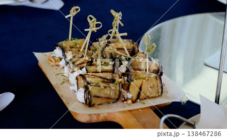 Grilled eggplant rolls stuffed with creamy cheese, toasted nuts, aromatic herbs, plated on rustic wooden board with garnishing skewers 138467286