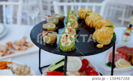 Slate round plate displaying elegant appetizers like pinwheel sandwiches, eclairs, and vegetable rolls, enhancing sophisticated catering presentation for festive gathering Slate round plate displaying elegant appetizers like pinwheel sandwiches, eclairs, and vegetable rolls, enhancing sophisticated catering presentation for festive gathering 138467288