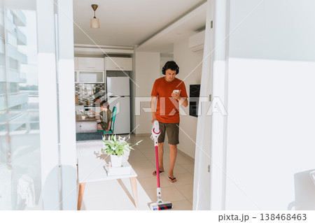 Man wireless vacuum cleaner headphones looking smartphone while spring-cleaning the house. Caucasian male in wireless earphones checking message in cellphone vacuum cleaning light cozy apartment Man wireless vacuum cleaner headphones looking smartphone while spring-cleaning the house. Caucasian male in wireless earphones checking message in cellphone vacuum cleaning light cozy apartment 138468463