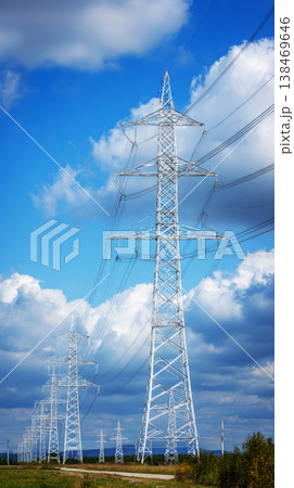 Tall electric power transmission towers stand along a road in the countryside. The lines connect the towers and stretch across the open land beneath a blue sky with clouds Tall electric power transmission towers stand along a road in the countryside. The lines connect the towers and stretch across the open land beneath a blue sky with clouds 138469646