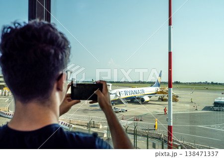 Passenger captures ryanair airplane at the gate with smartphone through terminal window, documenting travel moment on tarmac before boarding and departure into blue sky Passenger captures ryanair airplane at the gate with smartphone through terminal window, documenting travel moment on tarmac before boarding and departure into blue sky 138471337