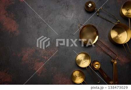 A gold-colored kitchen utensil set includes measuring cups of varying sizes, a sieve, spoons, and a tea strainer. Top view. A gold-colored kitchen utensil set includes measuring cups of varying sizes, a sieve, spoons, and a tea strainer. Top view. 138473029