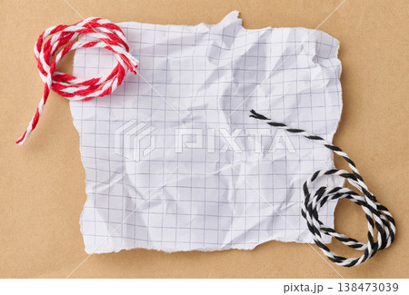 A crumpled, torn sheet of paper from a squared notebook lies on a brown background next to skeins of red, white, and black and white twine. 138473039