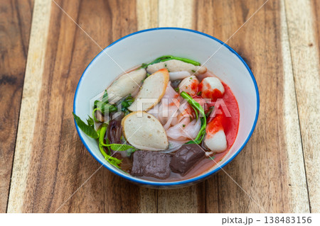 Thai noodle soup with fish balls herbs and vegetables in clear broth served in bowl on wooden table traditional street food fresh and appetizing meal Thai noodle soup with fish balls herbs and vegetables in clear broth served in bowl on wooden table traditional street food fresh and appetizing meal 138483156