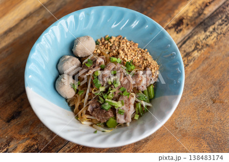 Thai dry noodle dish with pork slices meatballs bean sprouts peanuts and greens in bowl on wooden table authentic street food cuisine savory and flavorful meal 138483174