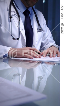 Medical professional wearing a white lab coat. Stethoscope. And tie. Sitting at a glass desk. Calculating paperwork. A laptop visible on the side 138483776