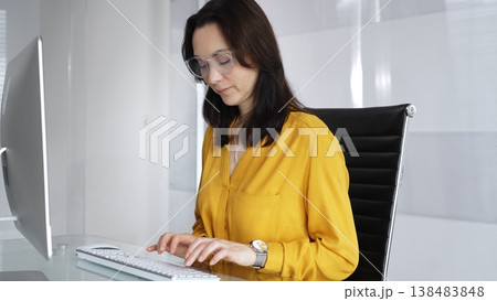 Businesswoman wearing yellow casual blouse and glasses is typing on laptop at office glass desk, analyzing data for business tasks. Audit in business Businesswoman wearing yellow casual blouse and glasses is typing on laptop at office glass desk, analyzing data for business tasks. Audit in business 138483848