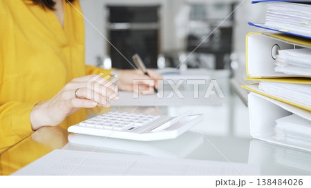 Woman in yellow blouse calculating finances at office desk, using calculator and writing, surrounded by documents and binders. Audit and taxes in business Woman in yellow blouse calculating finances at office desk, using calculator and writing, surrounded by documents and binders. Audit and taxes in business 138484026