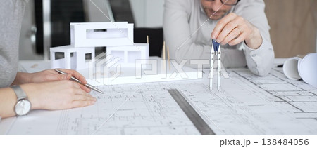 Architects collaborating on architectural design, discussing plans and blueprints with a scale model of a modern house on a desk during a business meeting, close up Architects collaborating on architectural design, discussing plans and blueprints with a scale model of a modern house on a desk during a business meeting, close up 138484056