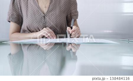 Businesswoman in brawn dotted blouse is signing a contract at a sleek glass desk in a modern office, showcasing professionalism and focus in her work environment, closeup. Business people concept Businesswoman in brawn dotted blouse is signing a contract at a sleek glass desk in a modern office, showcasing professionalism and focus in her work environment, closeup. Business people concept 138484116