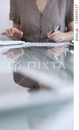 Businesswoman accountant, wearing brawn dotted blouse, is using a calculator at sleek glass desk in a contemporary office, vertical view. Taxes, audit in business 138484117