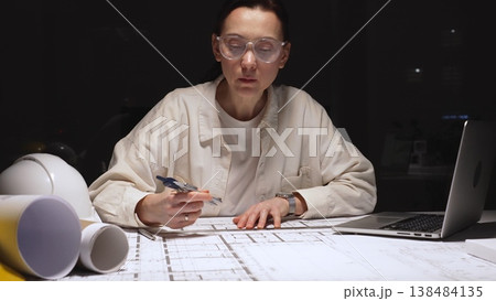 Female architect working late on detailed building blueprints using ruler and pencil, modern engineer designing construction plans at desk with laptop and hardhat 138484135