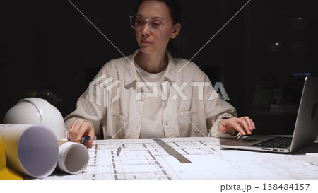 Female architect working late on detailed building blueprints using ruler and pencil, modern engineer designing construction plans at desk with laptop and hardhat 138484157