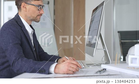 Architect working on computer in design office, planning and developing building projects with blueprints and models. Business and architecture concept 138484253
