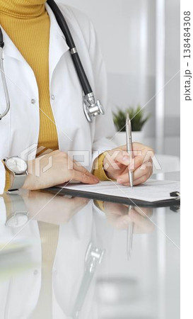 Doctor wearing stethoscope and yellow sweater under white lab coat, making notes on a clipboard in a bright medical office. Medicine, healthcare and science concept Doctor wearing stethoscope and yellow sweater under white lab coat, making notes on a clipboard in a bright medical office. Medicine, healthcare and science concept 138484308