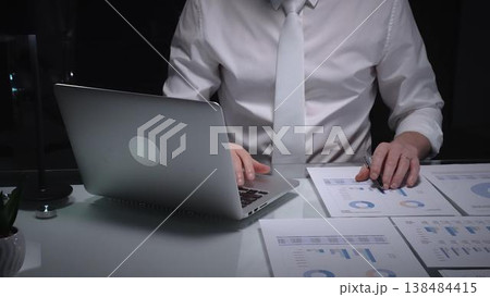 Business person working late overtime in dark office, typing on laptop with financial documents spread on desk. Audit and taxes Business person working late overtime in dark office, typing on laptop with financial documents spread on desk. Audit and taxes 138484415