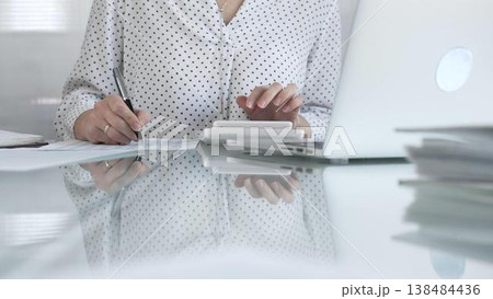 Focused businesswoman at a sleek glass desk in a contemporary office, using a calculator to crunch numbers and jotting down meticulous notes. Taxes, audit in business 138484436