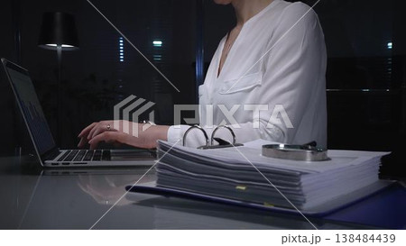 Businesswomen working overtime in a dark office, typing on laptop with financial charts and data reports on desk at night. Audit and taxes in business 138484439