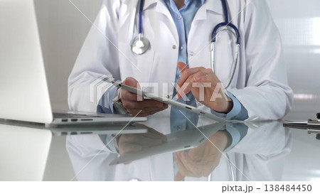 Doctor woman, wearing a blue blouse under a medical coat, is using digital tablet in modern healthcare setting, interacting with patient data, close-up. Medicine concept Doctor woman, wearing a blue blouse under a medical coat, is using digital tablet in modern healthcare setting, interacting with patient data, close-up. Medicine concept 138484450