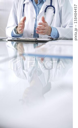 Unknown doctor woman, wearing a stethoscope, blue blouse under a lab coat, is gesturing with hands overs a glass desk with a laptop in clinic office, close up vertical view. Medicine and science Unknown doctor woman, wearing a stethoscope, blue blouse under a lab coat, is gesturing with hands overs a glass desk with a laptop in clinic office, close up vertical view. Medicine and science 138484457