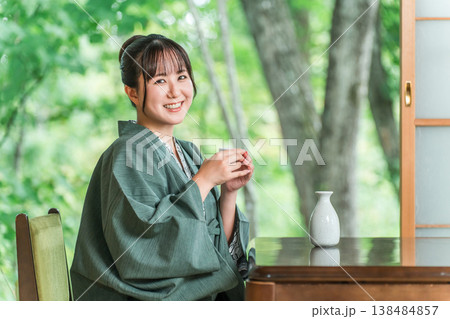 旅館の客室の縁側で日本酒を飲みながらくつろぐ浴衣羽織姿の若いアジア人女性 138484857