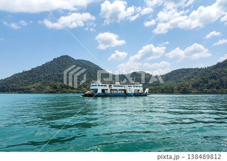 Large car ferry transporting tourists across the ocean to tropical island with mountain background. Large car ferry transporting tourists across the ocean to tropical island with mountain background. 138489812