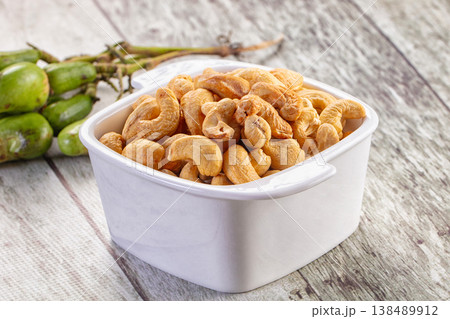 Roasted cashew nuts in a brown bowl with a cluster of raw green cashew fruit on a rustic wooden table. Roasted cashew nuts in a brown bowl with a cluster of raw green cashew fruit on a rustic wooden table. 138489912