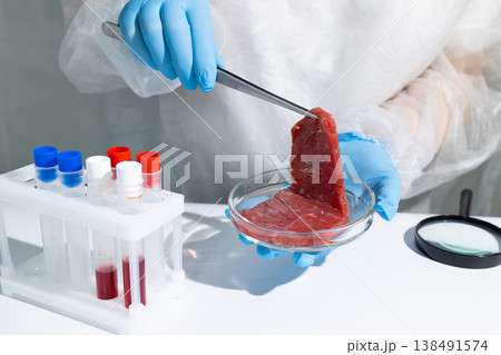 Scientist in protective gear examining lab-grown meat sample with tweezers over petri dish. In vitro cultivation process in biotechnology lab. Sustainable artificial protein production for hunger Scientist in protective gear examining lab-grown meat sample with tweezers over petri dish. In vitro cultivation process in biotechnology lab. Sustainable artificial protein production for hunger 138491574