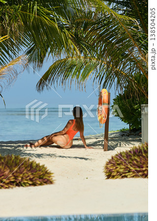 A beautiful tanned girl with an athletic physique in a swimsuit looks at the sea through palm trees on the personal beach of the villa on a sunny day in the Maldives, she is happy and posing A beautiful tanned girl with an athletic physique in a swimsuit looks at the sea through palm trees on the personal beach of the villa on a sunny day in the Maldives, she is happy and posing 138504205