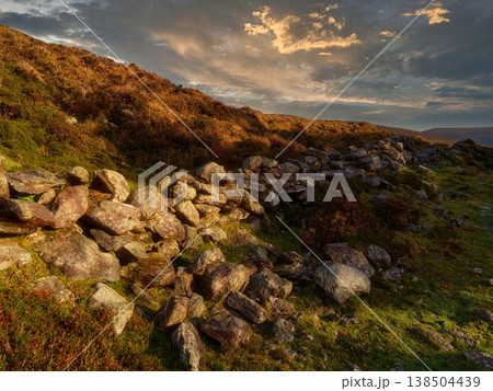 An old stone wall sits on a grassy hillside under a cloudy sky during sunset. 138504439