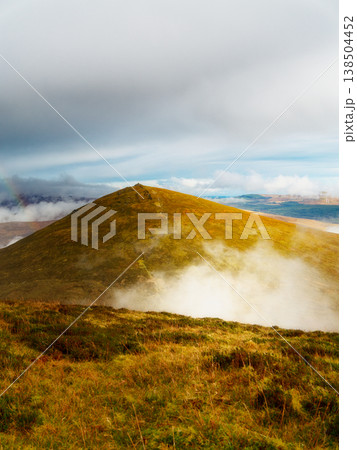 Golden sun shines on a vast mountain, its expansive slopes covered in rich grass and rising mist. The cloudy sky shows glimpses of blue, suggesting changing weather over this grand natural landscape. 138504452