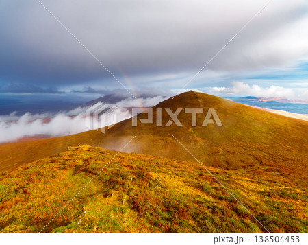 Golden green mountain slopes leading to distant peaks, a subtle rainbow visible through dramatic clouds. 138504453