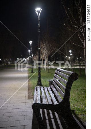 Empty park bench resting beside a paved pathway with modern streetlights creating a peaceful ambiance, offering a quiet space for contemplation or relaxation outdoors 138504635