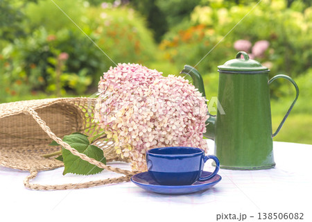 Summer leisure activity still life; blue cup of coffee, green vintage coffee pot, pnk hydrangea flower and wicker bag on a table in a summer blooming garden Summer leisure activity still life; blue cup of coffee, green vintage coffee pot, pnk hydrangea flower and wicker bag on a table in a summer blooming garden 138506082