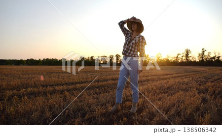 Adult agronomist in straw hat exploring barley on meadow at sunset. Female farmer examining ripe wheat at field on dusk. Beautiful scenic landscape. Concept of agriculture and agronomy business Adult agronomist in straw hat exploring barley on meadow at sunset. Female farmer examining ripe wheat at field on dusk. Beautiful scenic landscape. Concept of agriculture and agronomy business 138506342
