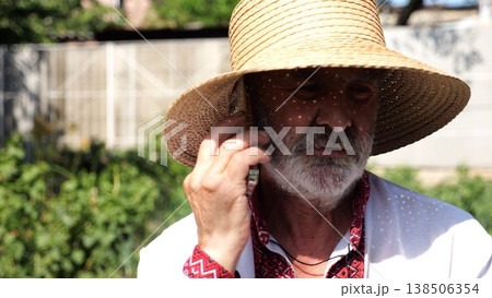 Older ukrainian man wearing in vyshyvanka talking on mobile phone at summer garden. Senior male in straw hat using cell phone outdoor. Concept of connection between tradition and modern life Older ukrainian man wearing in vyshyvanka talking on mobile phone at summer garden. Senior male in straw hat using cell phone outdoor. Concept of connection between tradition and modern life 138506354