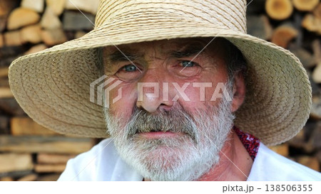 Portrait of senior ukrainian man in straw hat against the background of woodshed. Elderly serious male wearing an embroidered shirt looking into camera outdoor. Cultural heritage and ethnic roots Portrait of senior ukrainian man in straw hat against the background of woodshed. Elderly serious male wearing an embroidered shirt looking into camera outdoor. Cultural heritage and ethnic roots 138506355