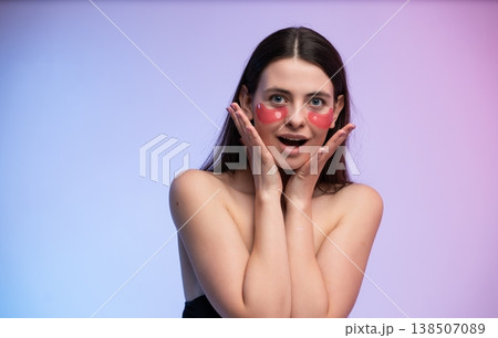 Young woman applying red hydrogel eye patches for skincare treatment on her face, experiencing self-care and beauty with a surprised expression and glowing skin Young woman applying red hydrogel eye patches for skincare treatment on her face, experiencing self-care and beauty with a surprised expression and glowing skin 138507089