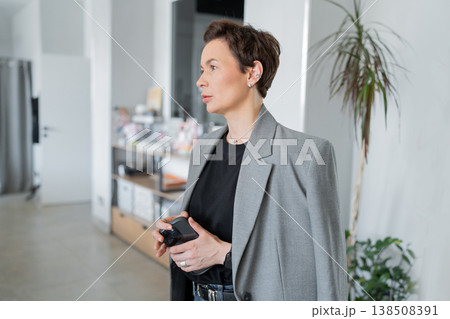 Businesswoman smiling and holding folder while talking to colleague in bright office with green plant. Smiling, talking, businesswoman, office, folder, friendly. Businesswoman smiling and holding folder while talking to colleague in bright office with green plant. Smiling, talking, businesswoman, office, folder, friendly. 138508391
