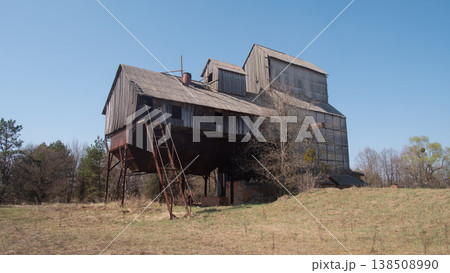 Abandoned Wooden Farmhouse In Field With Rusted External Stairs, Boarded Windows, Overgrown Grass, Surrounding Pine Trees Under Clear Blue Sky, Atmosphere Of Isolation And Quiet, Visible 138508990
