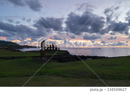 Majestic moai statue silhouette stands quietly on green easter island coast during peaceful sunset over ocean. serene rapa nui landscape features heavy dark cloud filling sky 138509627