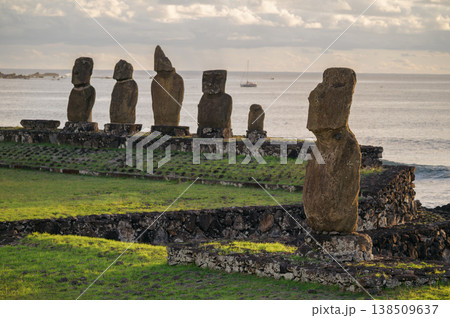 Majestic moai statue stands stoic on grassy shore of rapa nui easter island looking over peaceful ocean water beside an ancient stone monument under grey cloudy sky 138509637