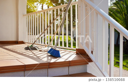 Brooms and mops leaning on white fence at hotel room terrace, morning sun shines on, blurred palm trees background - cleaning in tourist resort concept 138510088