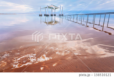 Calm morning at Ein Bokek Dead Sea beach, pink clouds reflection over water surface near steel rails leading to sun shade shelter, salt crystals on wet sand beach Calm morning at Ein Bokek Dead Sea beach, pink clouds reflection over water surface near steel rails leading to sun shade shelter, salt crystals on wet sand beach 138510121