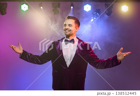 Happy presenter in bow tie on stage under vibrant lights with open arms wide Happy presenter in bow tie on stage under vibrant lights with open arms wide 138512445