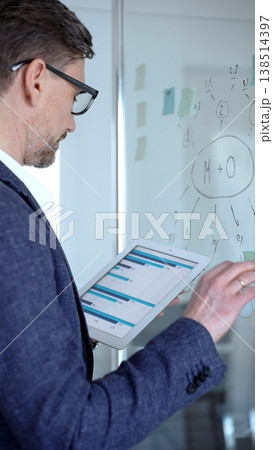 Professional businessman in modern office environment developing new business strategies. Analyzing data on a digital tablet while brainstorming on a glass wall or whiteboard Professional businessman in modern office environment developing new business strategies. Analyzing data on a digital tablet while brainstorming on a glass wall or whiteboard 138514397