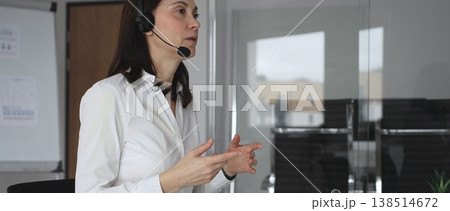 Businesswoman in a white shirt and headset leading a video call from a modern office, presenting with a tablet and gesturing as she discusses strategy and trains remote colleagues Businesswoman in a white shirt and headset leading a video call from a modern office, presenting with a tablet and gesturing as she discusses strategy and trains remote colleagues 138514672