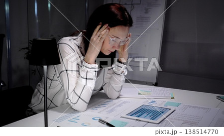 Businesswoman clutching her head in a dim office, surrounded by paperwork and a tablet while working late, showing exhaustion, headache and overwhelming professional stress Businesswoman clutching her head in a dim office, surrounded by paperwork and a tablet while working late, showing exhaustion, headache and overwhelming professional stress 138514770